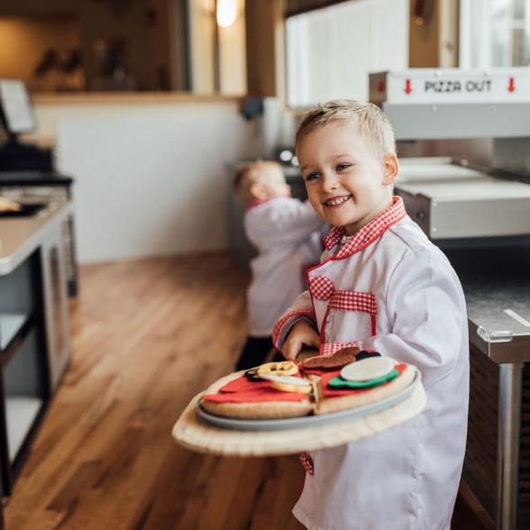 Young boy dressed as a chef holding a pretend pizza ready to go into a play oven.